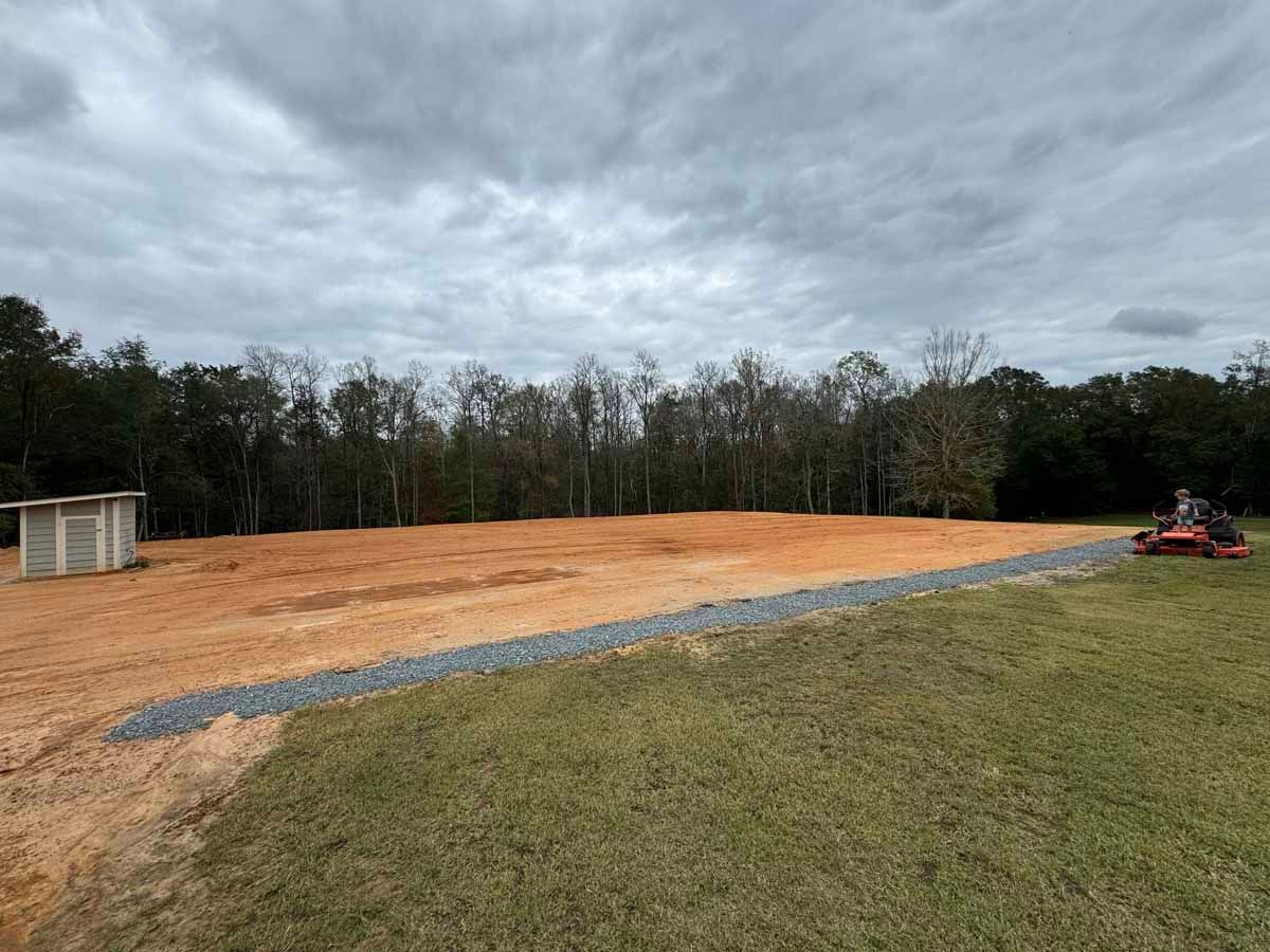 Cleared, leveled area with gravel border, small shed, and lawnmower on grass. Overcast sky, woods in the background.