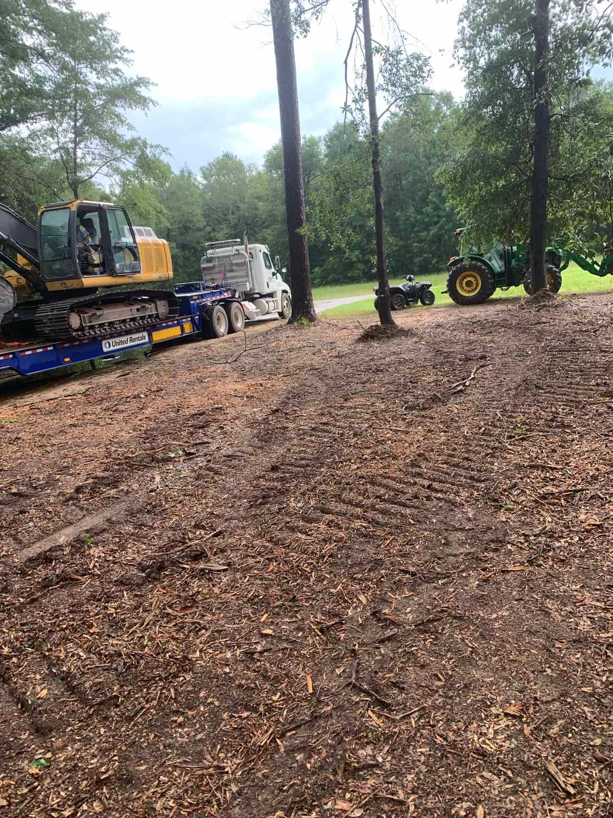 An excavator on a flatbed truck, tractor, and ATV in a muddy, wooded area.