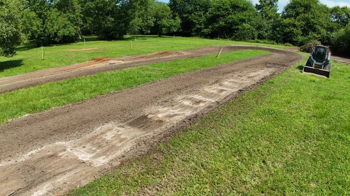 A small tractor on a grassy field has cleared two paths in the soil; trees are in the background.