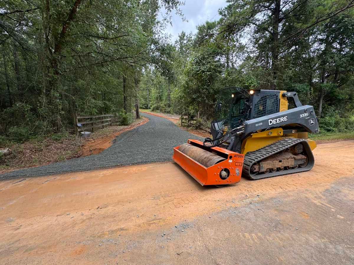 Skid steer compacting gravel on a dirt road through a wooded area.