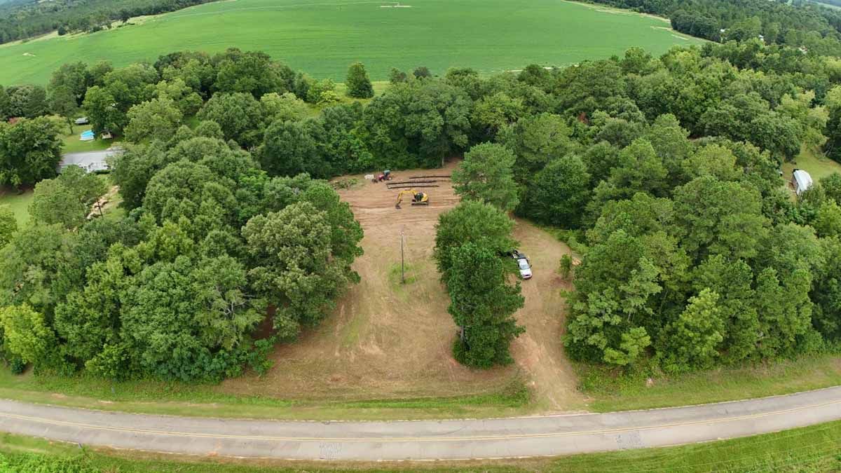 Aerial view of a cleared area with trees, a dirt road, and a green field in the background.