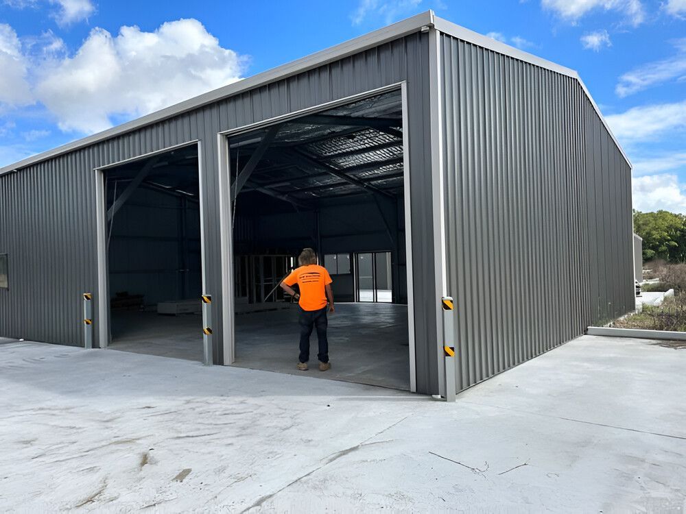A Man In An Orange Shirt Is Standing In Front Of A Large Metal Building — Mbah Metal Roofing In Murwillumbah, NSW