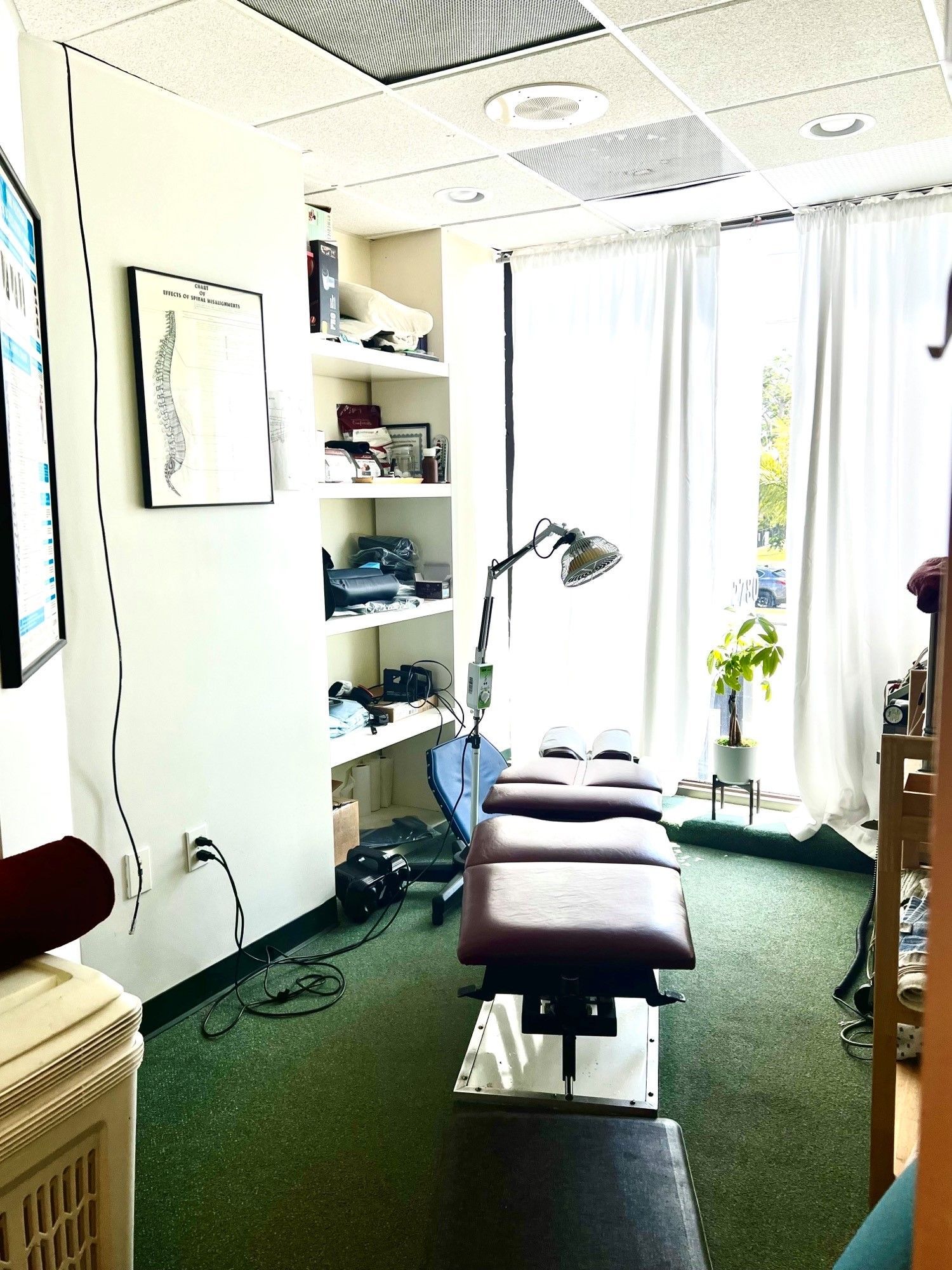 A chiropractic treatment room with a central adjustment table, an examination lamp, wall-mounted spine chart, and shelving.