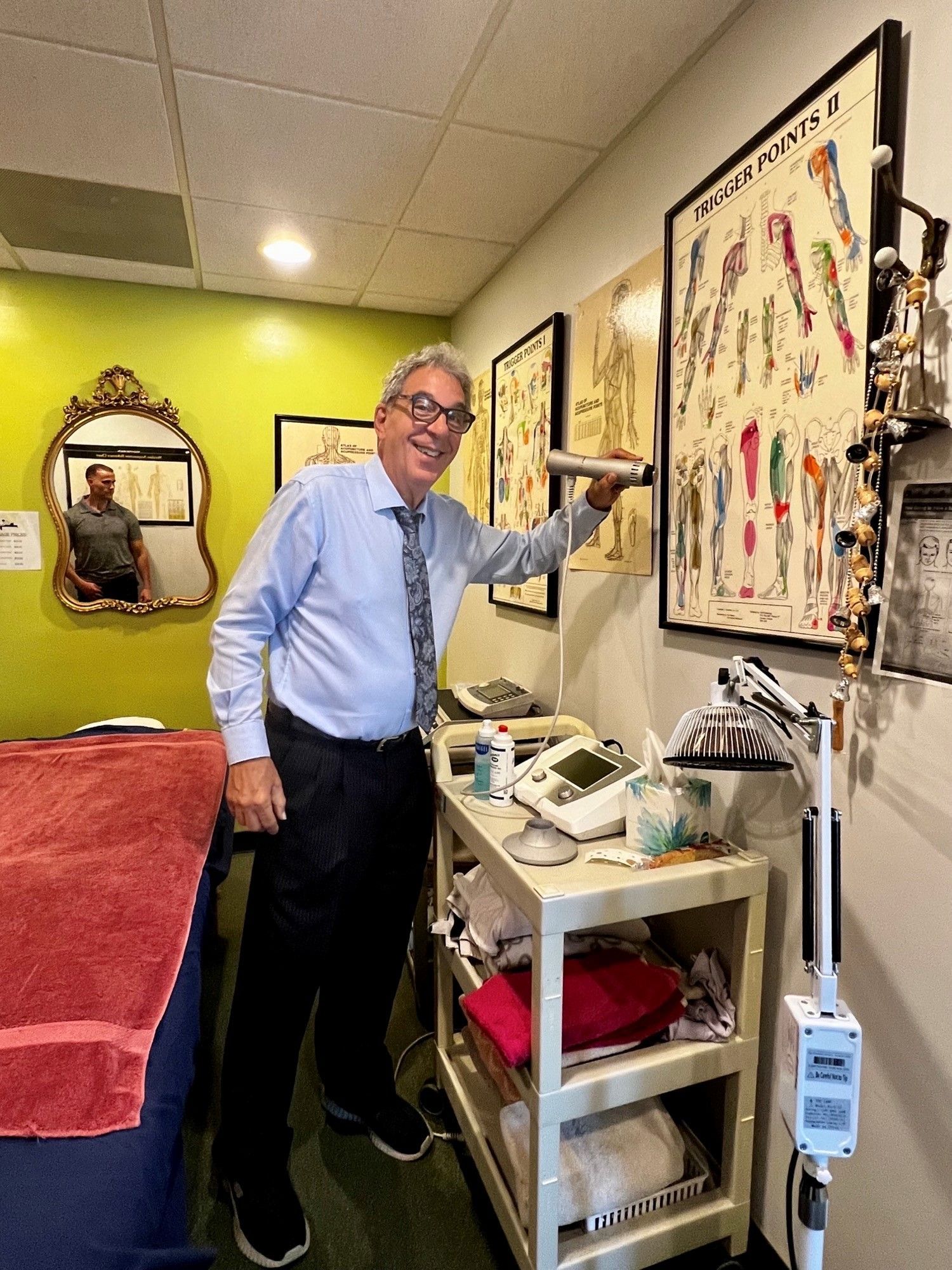 A professional in a blue shirt and tie points to an anatomical wall chart in a treatment room with a massage table.