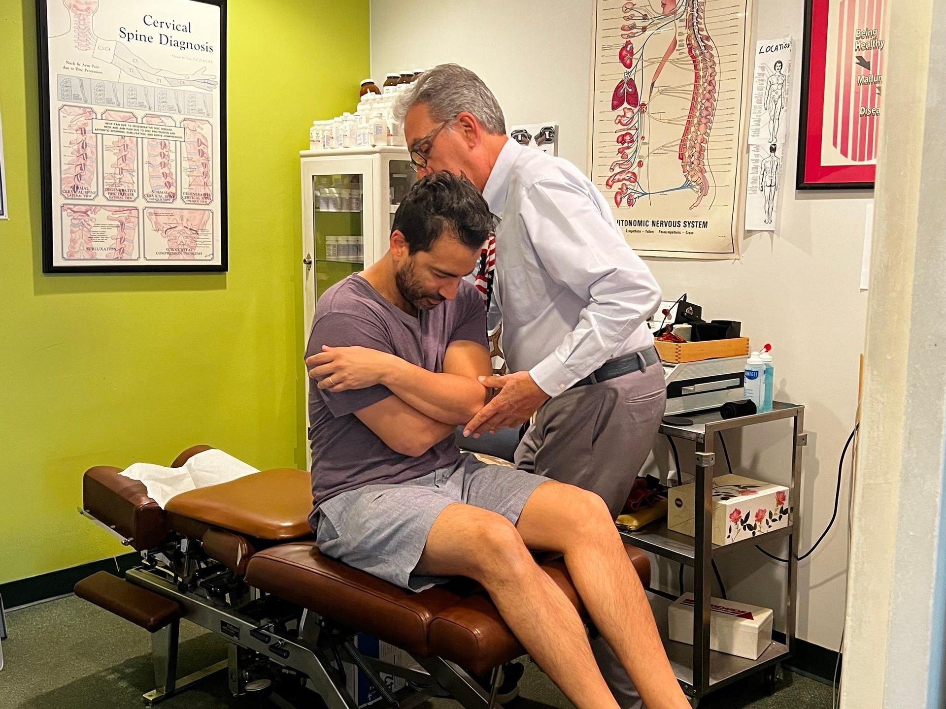A chiropractor adjusts a patient who is seated on a treatment table with arms crossed in a clinical office.