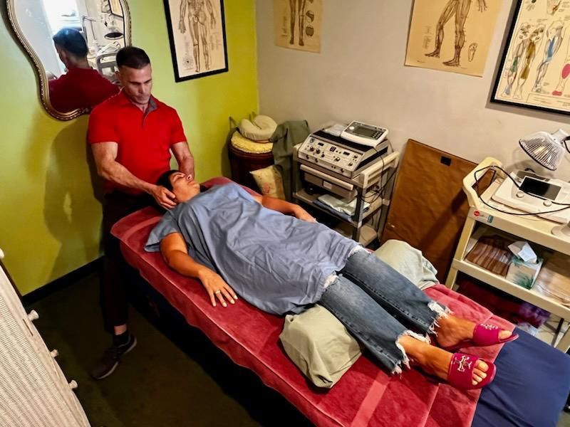 A person in a red shirt performs a neck adjustment on a person lying on a massage table in a clinical setting.
