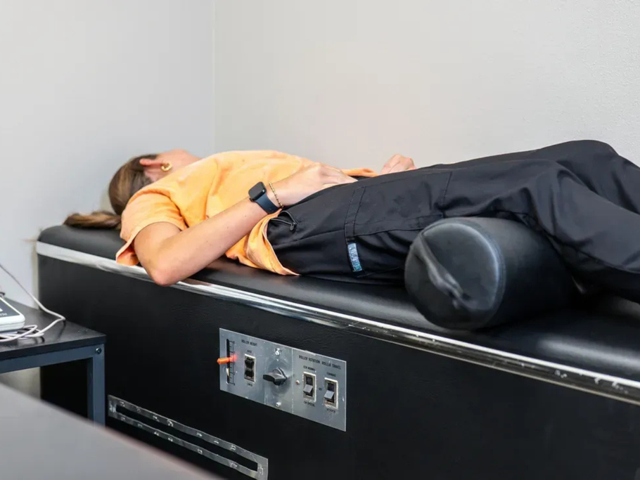 A person lying on their back on a treatment table with a bolster under their knees in a clinical setting.