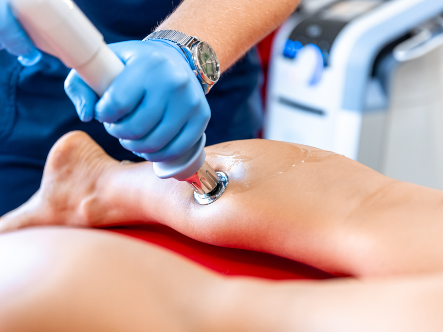 A person wearing blue medical gloves uses a laser therapy device on a client’s lower leg, which is resting on a red table.