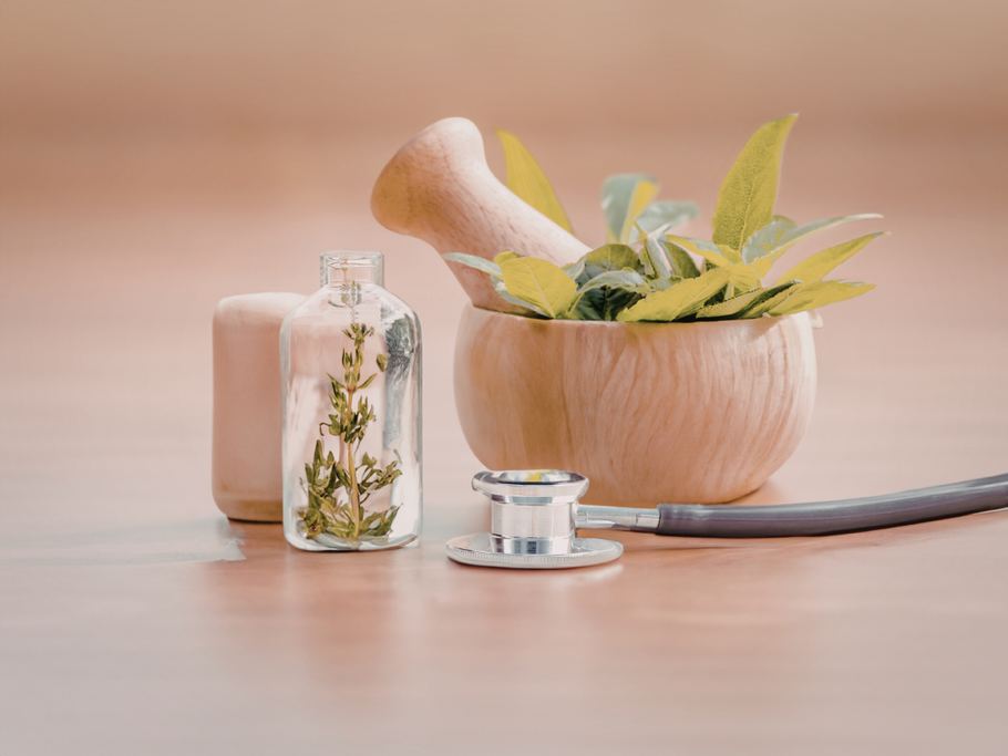 A mortar and pestle with green herbs, a small glass vial with dried plants, and a stethoscope on a wooden surface.
