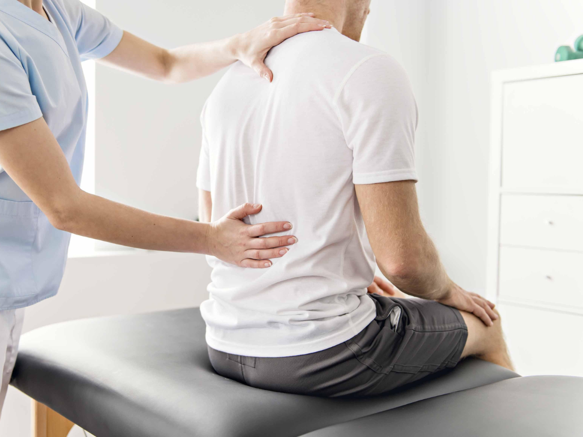 A healthcare professional in light blue scrubs performs a physical examination on a patient's back in a clinical setting.