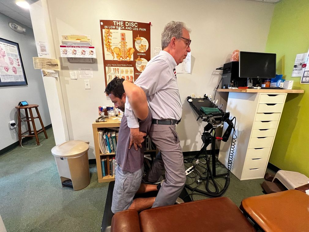 A chiropractor in a medical office performs a spinal adjustment on a patient sitting on an exercise bike.