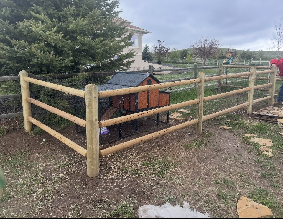 Wooden fenced enclosure around a small black-and-orange garden structure in a grassy yard under a cloudy sky