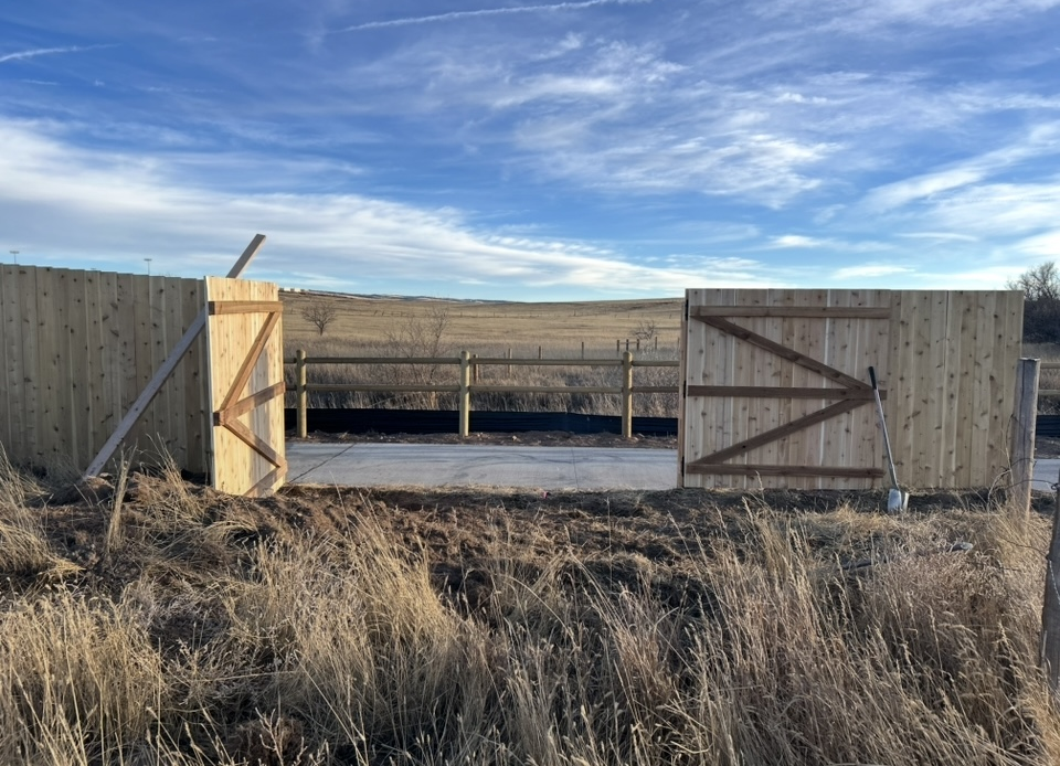 Open wooden gate in a dry field under a blue sky, with a fenced structure beyond.
