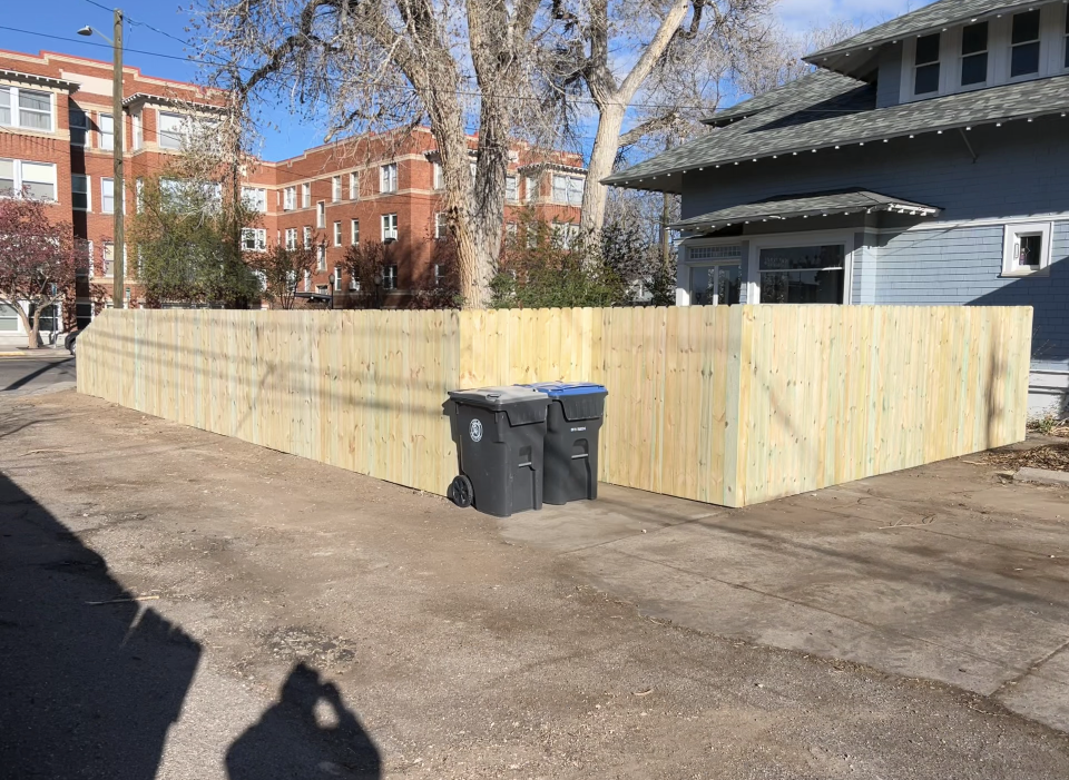 Temporary wooden fence enclosing a corner lot with two trash bins in front of apartment buildings