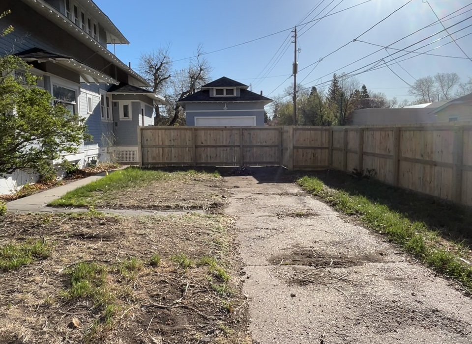 Backyard with patchy grass, gravel path, wooden fence, and houses under a clear sky