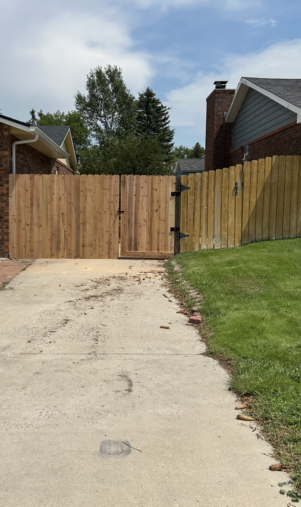 Wooden driveway gate beside a concrete path, with a grassy yard and houses under a cloudy sky.