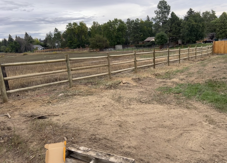 Dirt yard with a wooden fence, open field, and trees under a cloudy sky