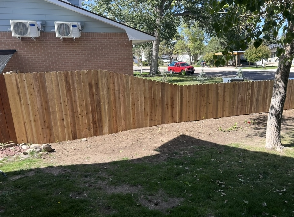 Wooden privacy fence beside a brick house in a sunny yard, with trees and a red vehicle in the background.