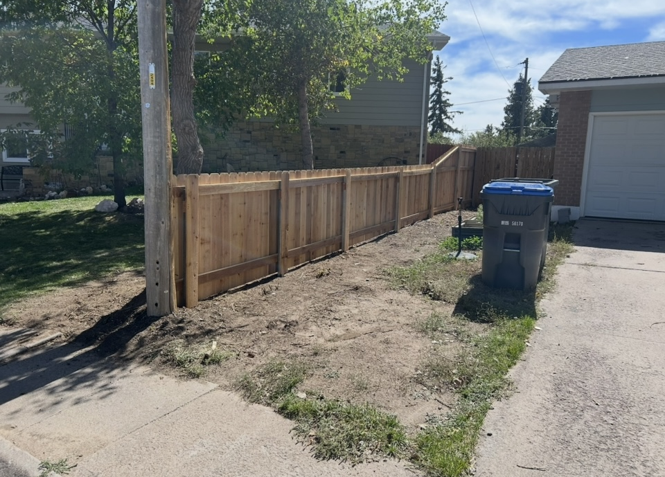 Wooden fence along a residential driveway beside a blue trash bin and garage, with trees and dry grass.