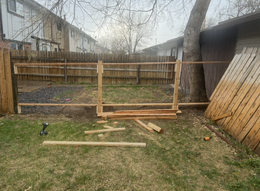 Wooden fence frame under construction in a backyard, with tools and lumber on the grass.