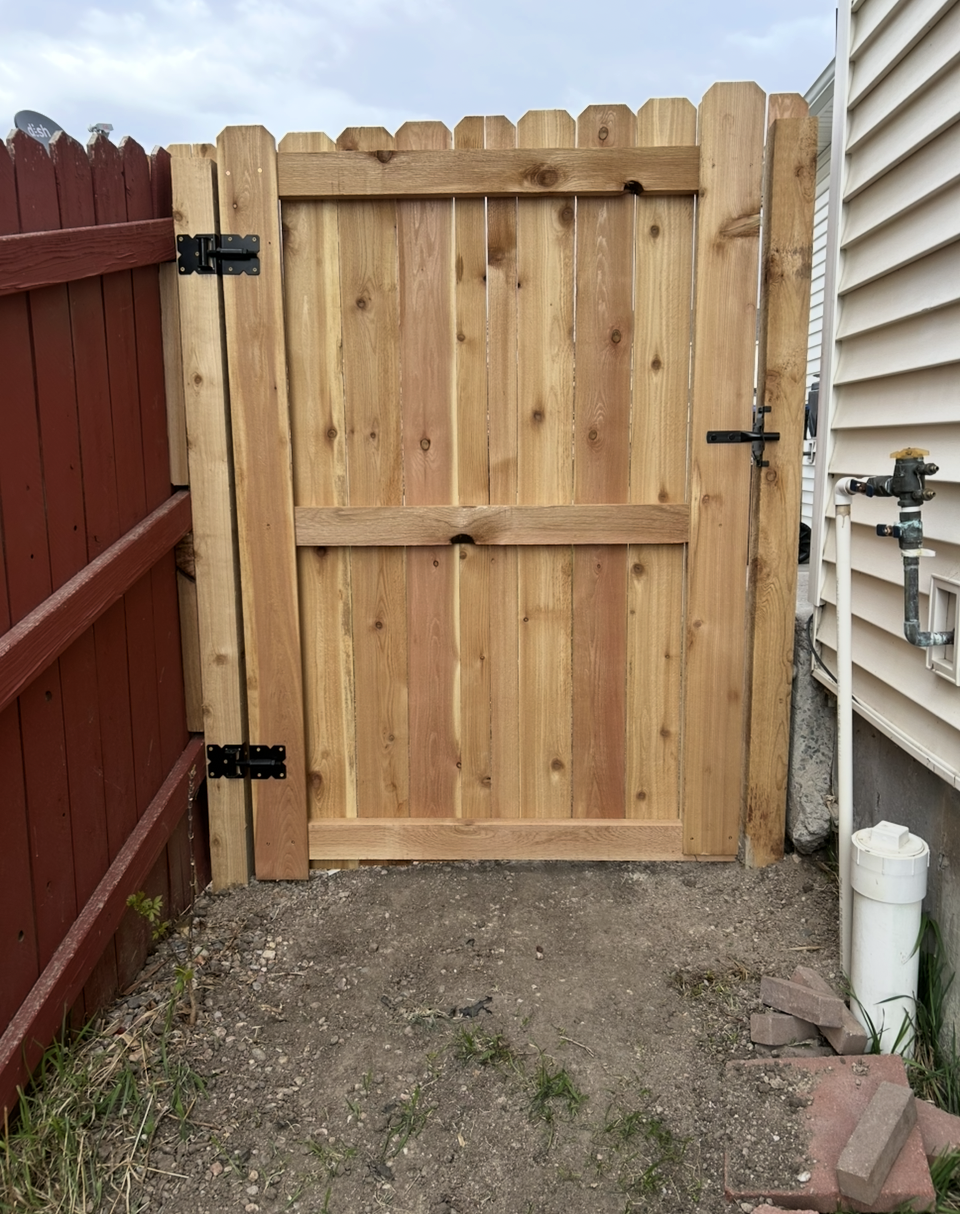 Wooden backyard gate between a red fence and house siding, with a gravel path in front.