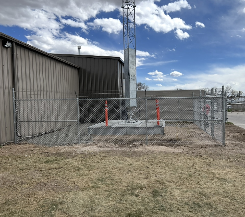 Chain-link fenced utility area beside a metal building, with red posts and a tall tower under a cloudy sky