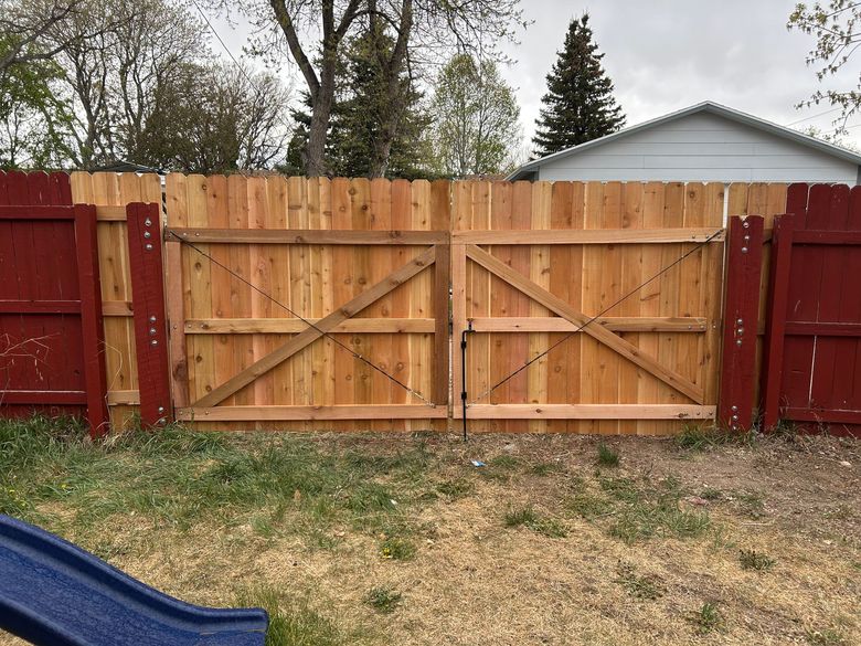 Wooden backyard gate with red posts and a fence, viewed over patchy grass under cloudy sky.