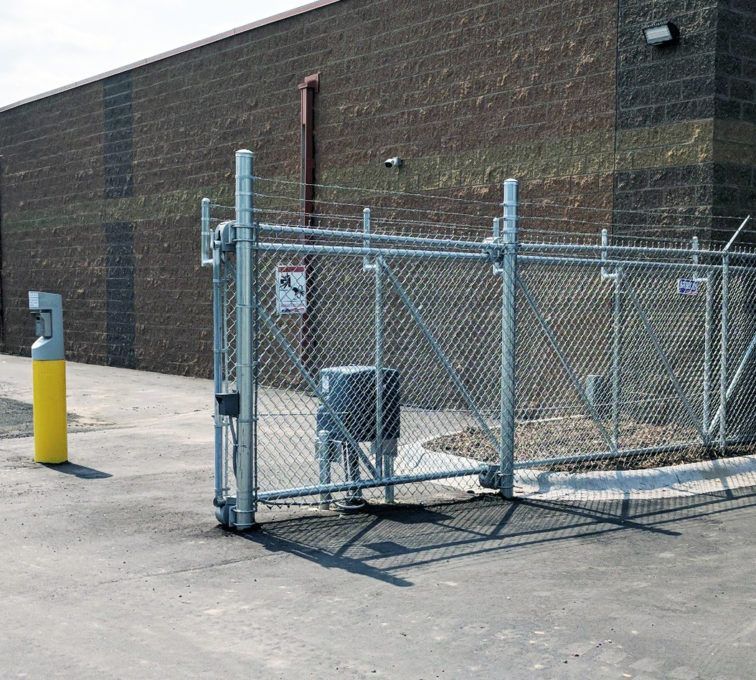 Chain-link fenced gate beside a brick building, with a yellow bollard on the pavement.