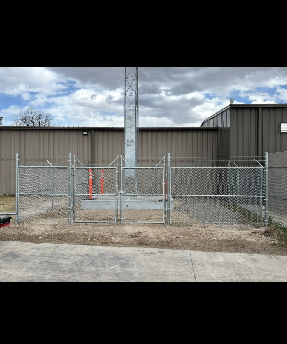 Chain-link fenced utility area beside a gray industrial building with a utility pole and orange cone.