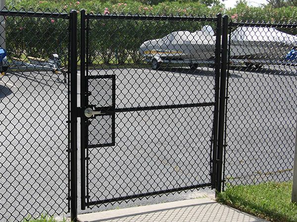 Black chain-link gate with lock on a paved driveway, with parked cars in the background.
