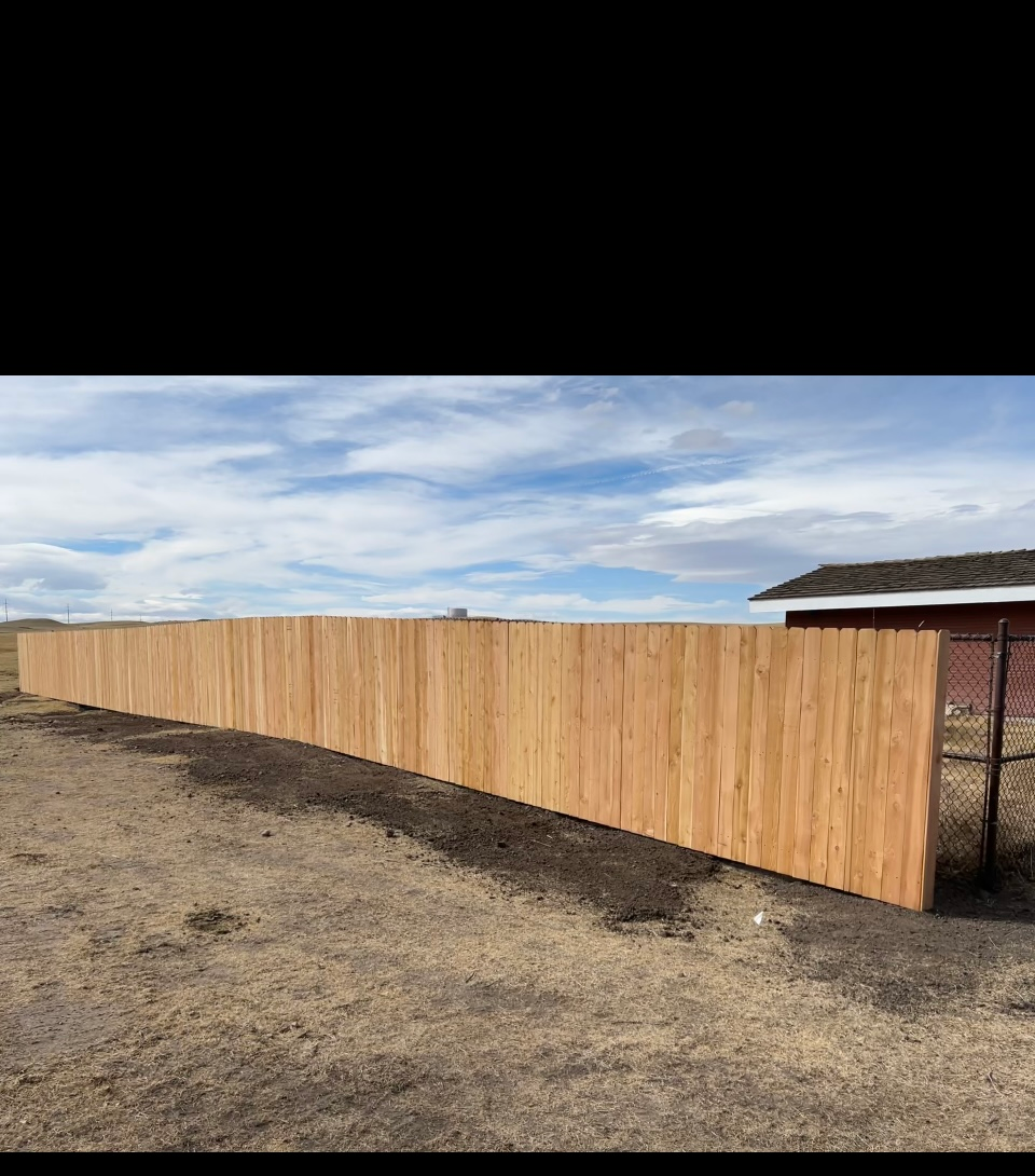 Long wooden fence on bare ground beside a building under a cloudy sky