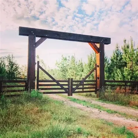 Wooden ranch gate with dark posts and crossbars on a dirt path, framed by green trees under a pink sky
