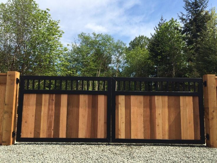 Wooden and black metal driveway gate in front of trees and blue sky