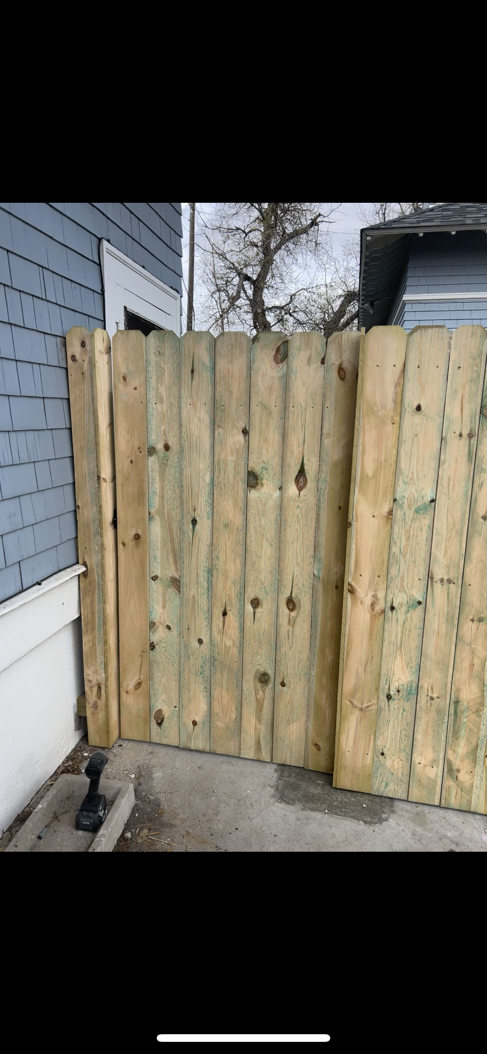 Wooden fence gate between a blue house and driveway, with a tree visible beyond it.