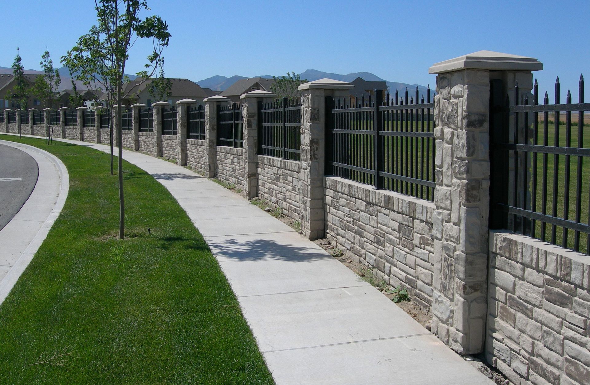 Curved sidewalk beside a stone-and-iron fence along a grassy roadside in a suburban neighborhood