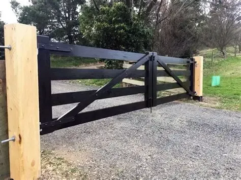 Black wooden farm gate with diagonal braces between wooden posts on a gravel driveway.