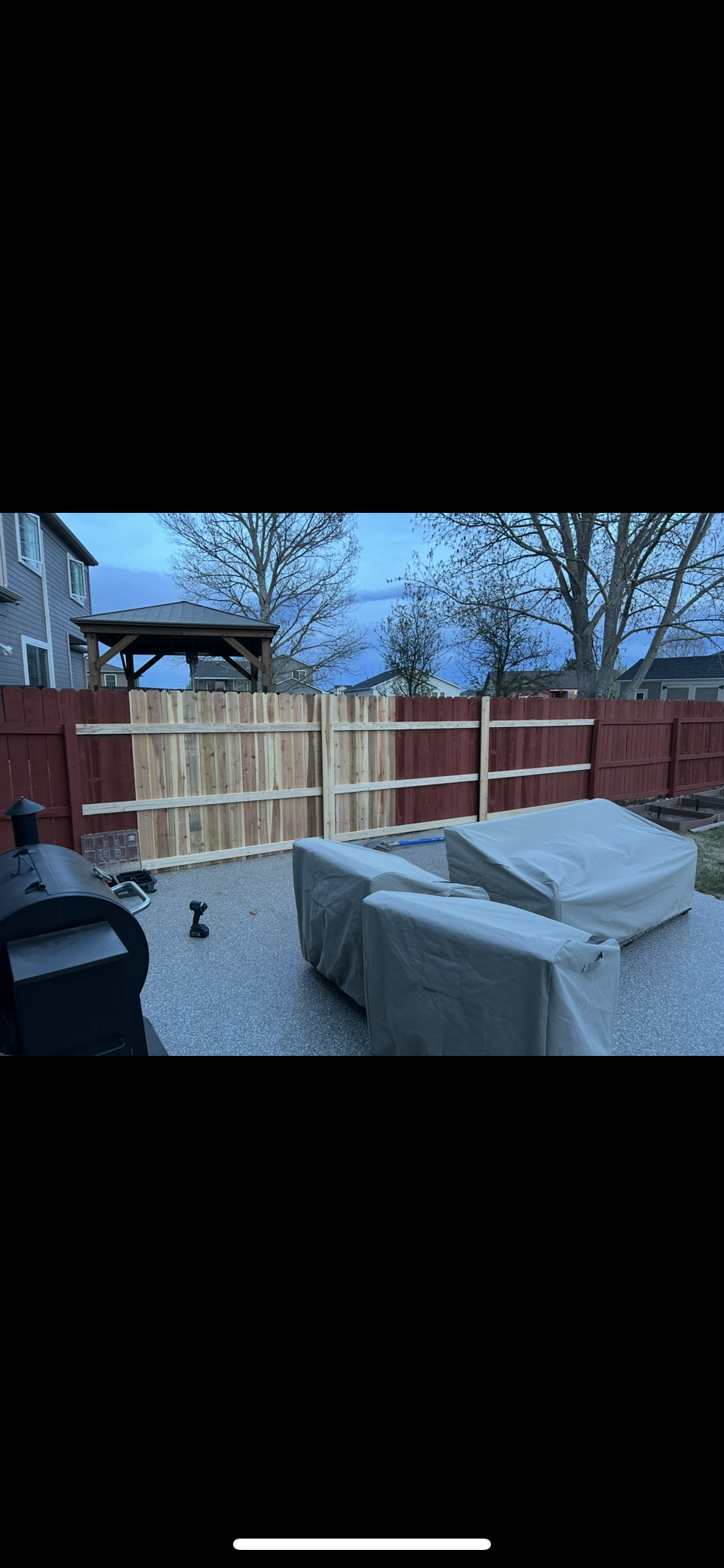 Backyard patio with covered furniture, wooden fence, gravel, and trees under a gray sky