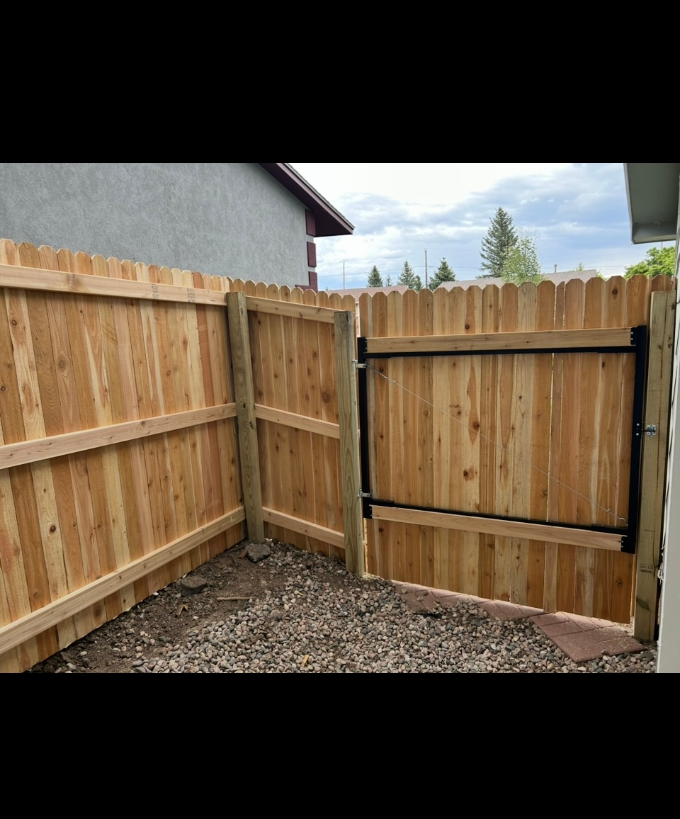 Wooden backyard fence corner with a closed gate and gravel ground beneath a cloudy sky