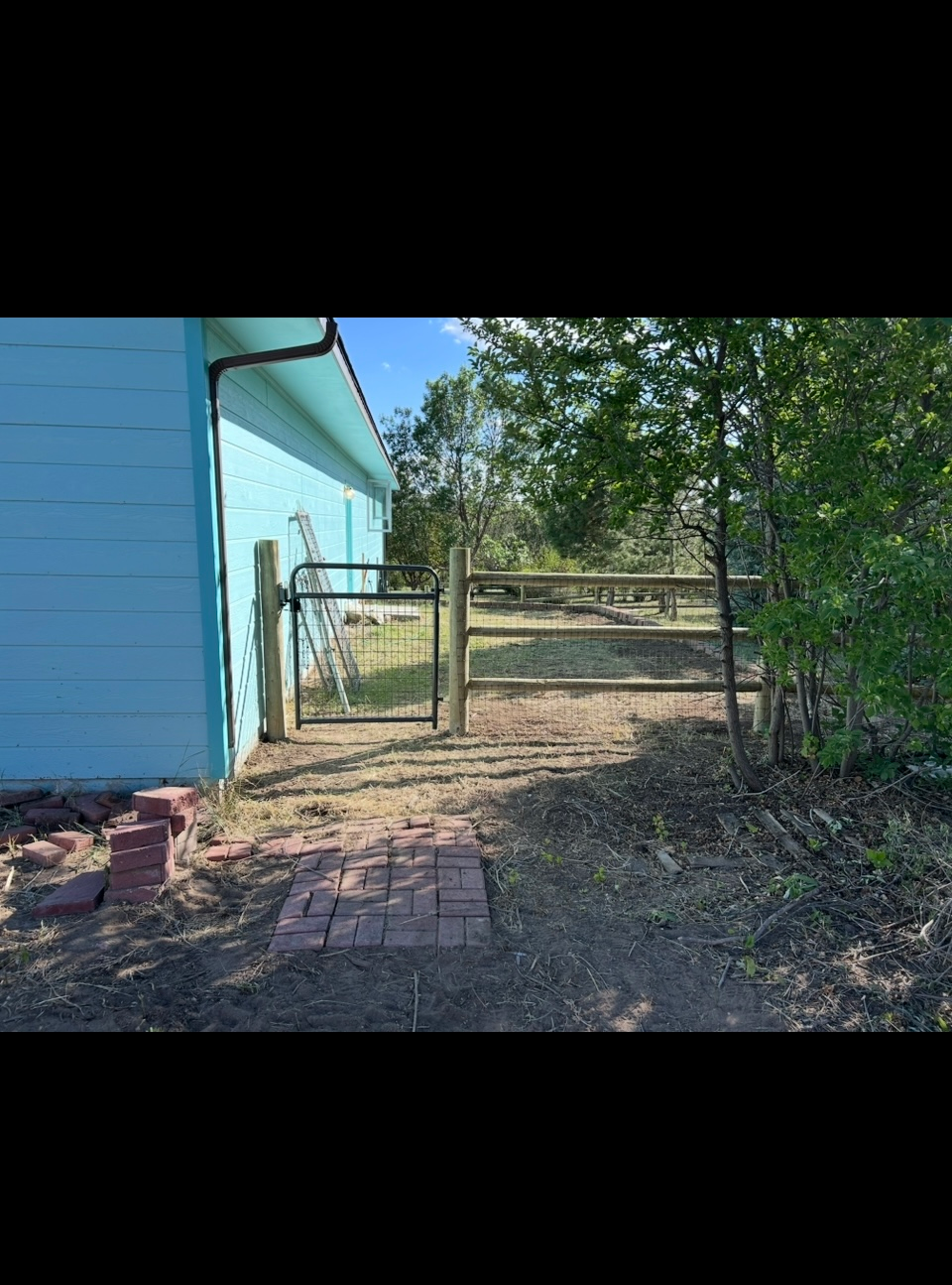 Blue house beside a fenced dirt path leading to a gate, with trees and dry ground nearby