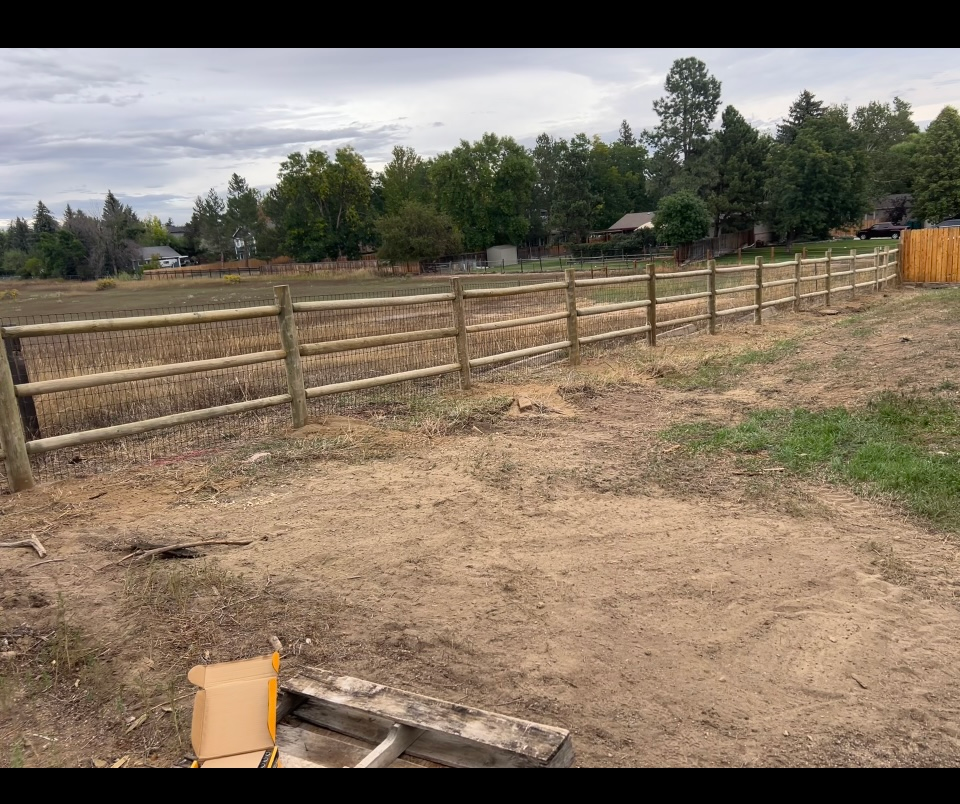 Wooden fence along a dirt field under cloudy skies, with trees in the background.