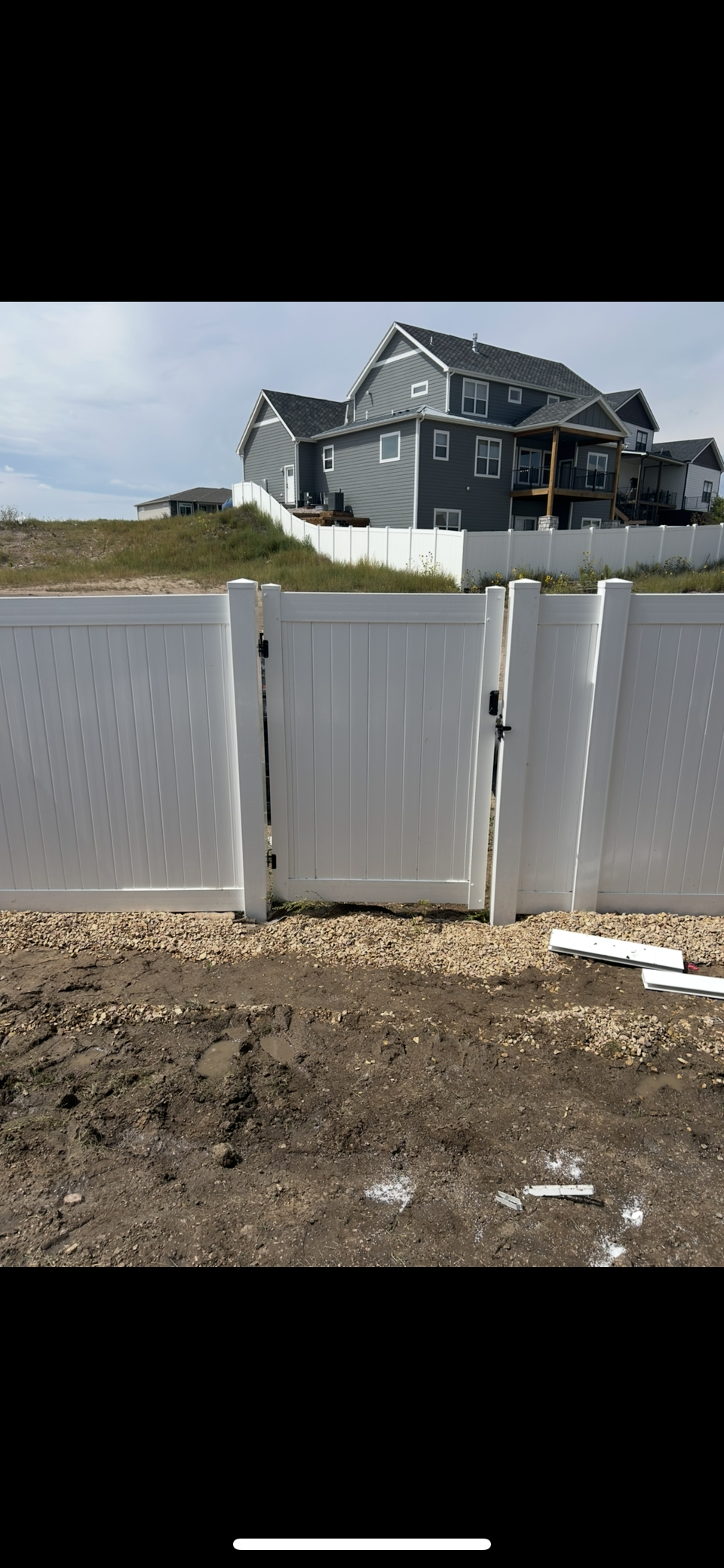 White fence in front of a damaged dark house on a dusty lot under a blue sky