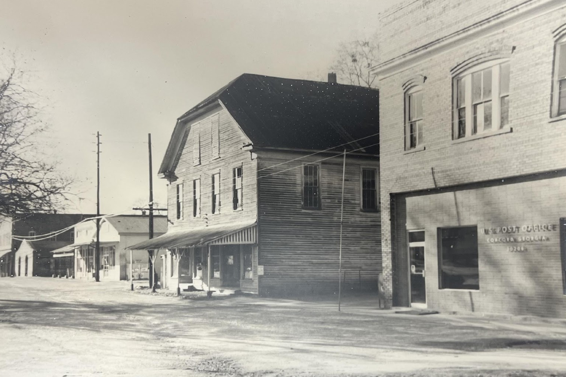 A black-and-white, historic street scene showing a row of small-town storefronts and buildings along a dirt road.