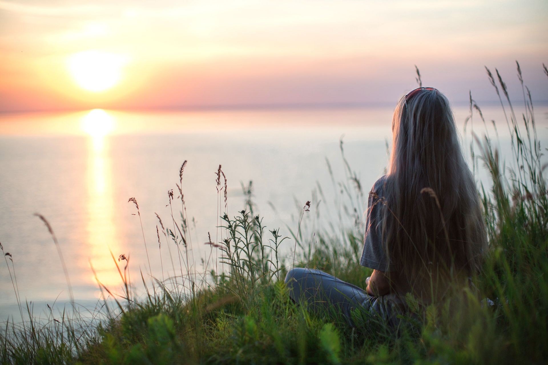 Person sitting in tall grass by a calm lake at sunset, facing the glowing horizon