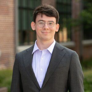 Man in glasses and suit smiling outdoors in front of a brick building.