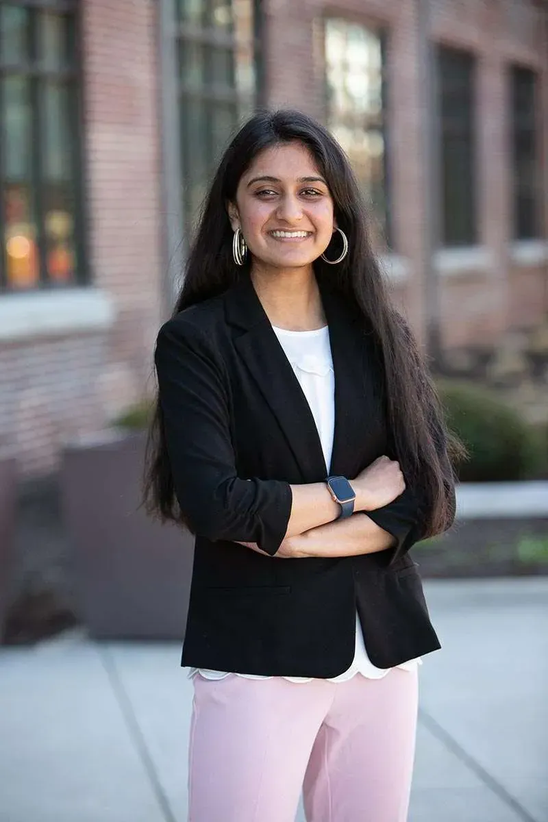 Woman in black blazer, pink pants smiles outdoors with arms crossed.