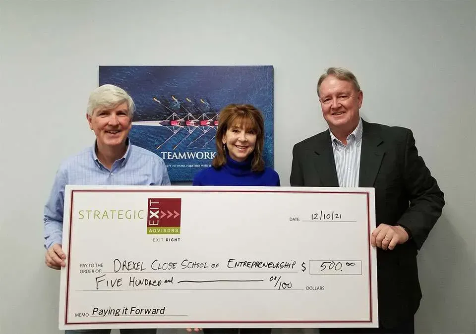 Three people holding a large check in an office, with a rowing team photo in the background. The check reads