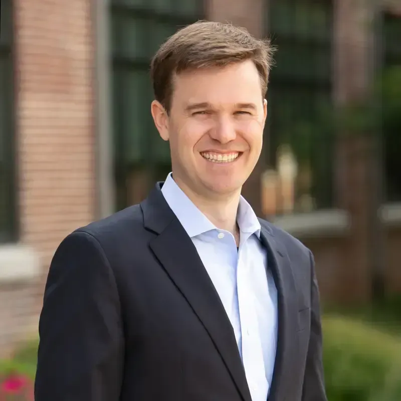 Man in dark suit and light blue shirt smiles outdoors in front of a brick building.