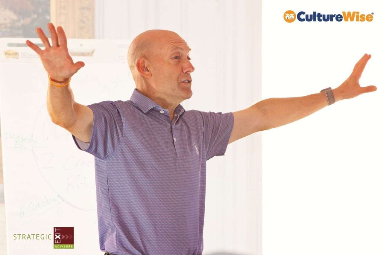 Man with arms outstretched, speaking. He wears a purple polo shirt, standing in front of a whiteboard.