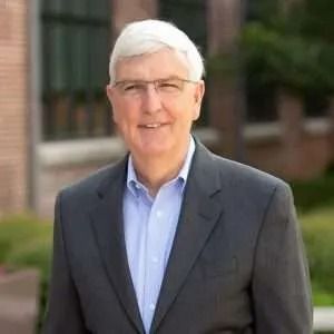 Man in a suit and glasses smiling outdoors in front of a building.