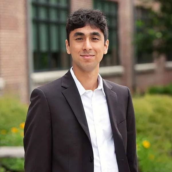 Man in black suit and white shirt smiles outdoors in front of a building.